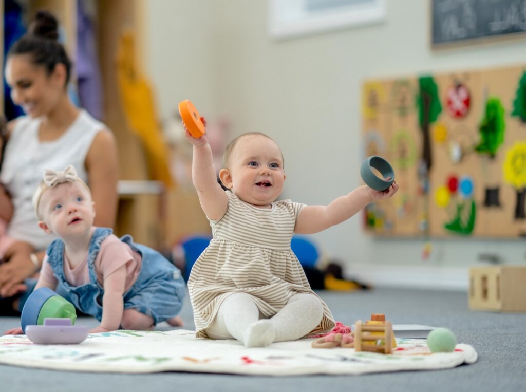 A group of babies is seen playing on the floor of their daycare classroom. They are each dressed casually and are playing with various toys.