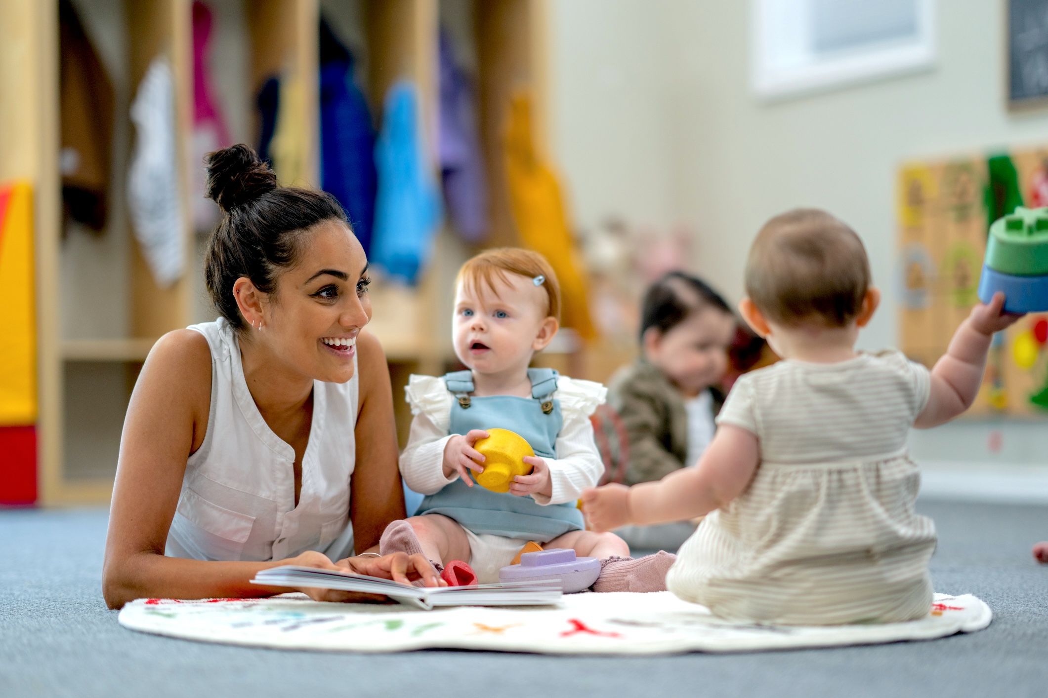 A small group of babies sit on the floor playing with various toys while their teacher supervises and engages them. They are each dressed casually and appear content.