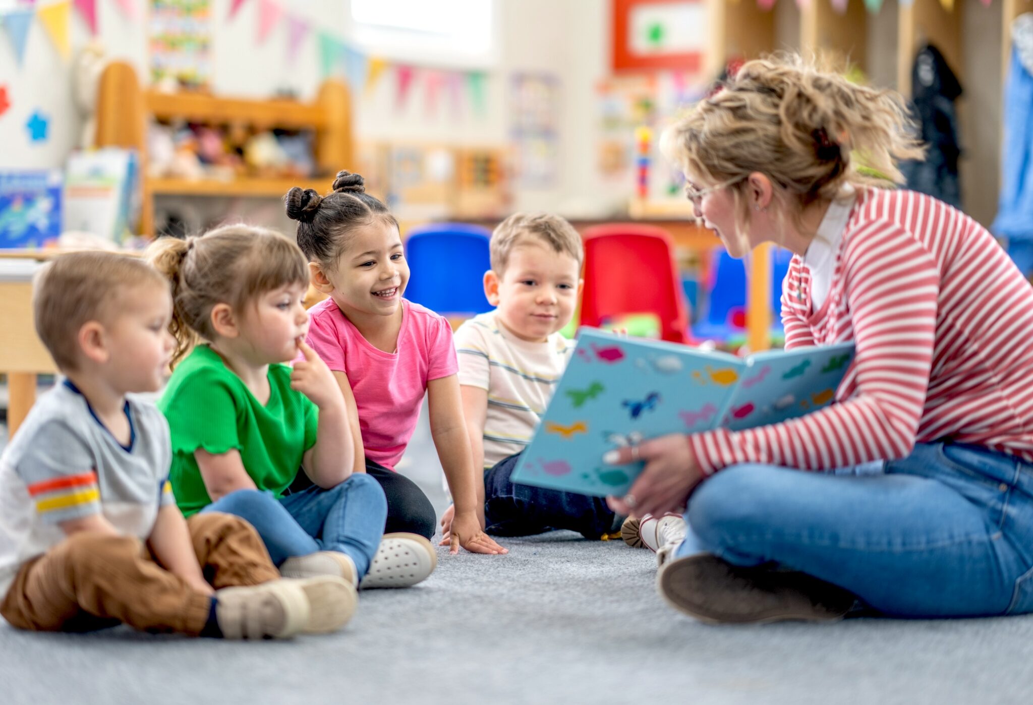 A preschool teacher sits on the floor of her classroom with a small group of students as she reads them a book. The children are each dressed casually and are focused on the story.
