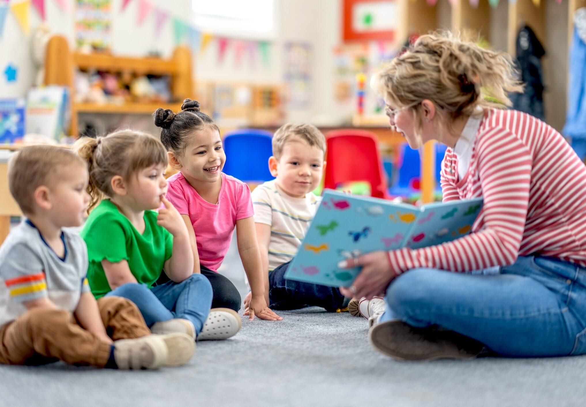 A preschool teacher sits on the floor of her classroom with a small group of students as she reads them a book. The children are each dressed casually and are focused on the story.
