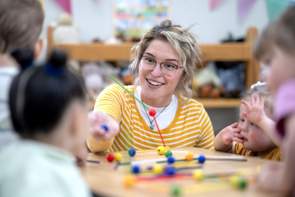 A teacher interacts with preschool children around a table filled with engaging learning tools, fostering creativity and education in a friendly classroom environment.
