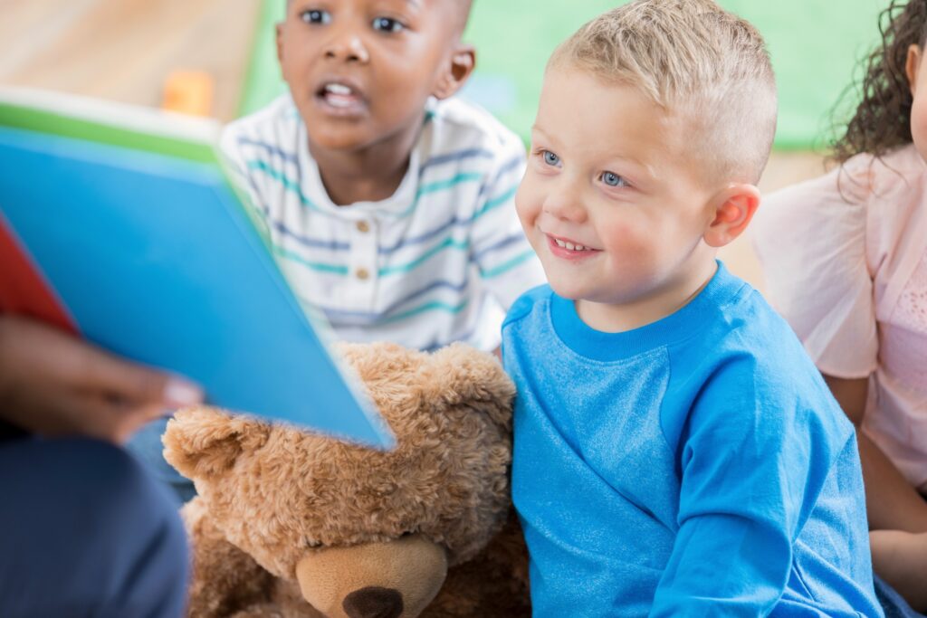 A smiling little boy sits on the floor of his classroom with his attentive classmates and listens to an unrecognizable day care worker during story time. He sits with his arm around a large teddy bear.