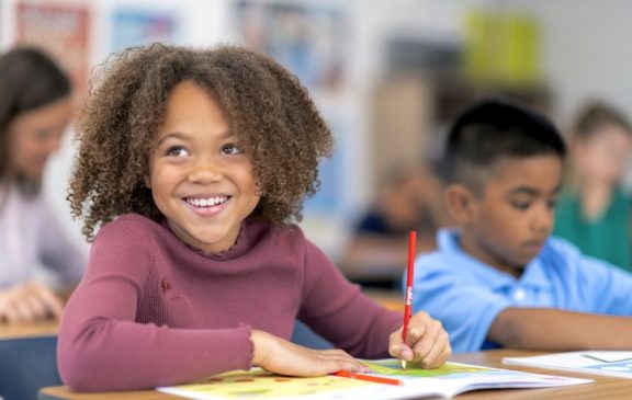 An elementary age girl is sitting at a desk in a classroom. The mixed-race African American child is working on an assignment in a notebook. She is smiling confidently. The image features a multi-ethnic group of students.