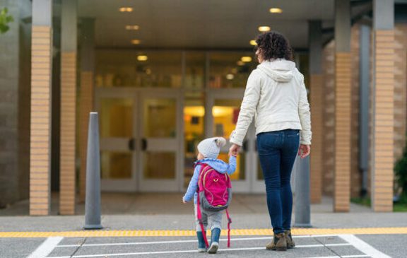 Rear view of a cheerful young mother holding her toddler daughter's hand while walking at crosswalk to the child's school.