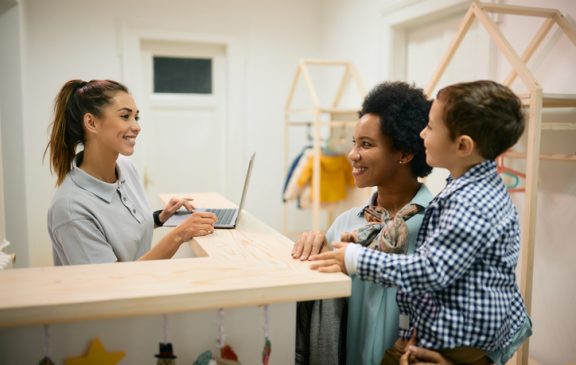 Happy kindergarten teacher communicating with African American mother and her son at reception desk.