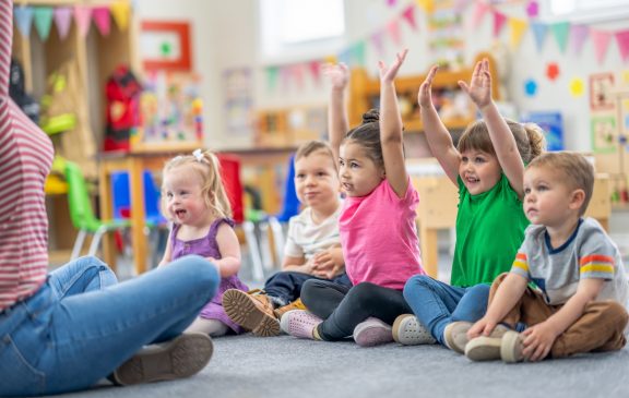 A preschool teacher sits on the floor of her classroom with her students in front of her as she leads them in a sing-along.  The children are dressed casually and smiling as they following along with the actions.