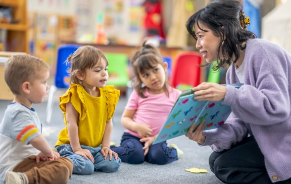 A preschool teacher sits on the floor of her classroom with a small group of students as she reads them a book.  The children are each dressed casually and are focused on the story.