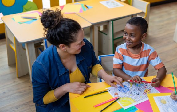 Latin American preschool teacher talking to a happy boy in the classroom while coloring - education concepts