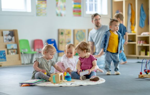 Young children playing together on a playmat in an organized daycare setting with vibrant colors and educational toys. A caregiver provides supervision, fostering a fun and safe learning environment.