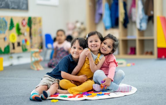 A small group of three kindergarten students huddle together closely on a rug as they pose for a portrait.  They are each dressed casually and smiling.
