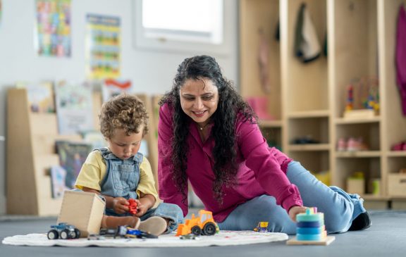 A caregiver and a child engaging in playful and educational activities together at a daycare. The image showcases a nurturing environment for childhood learning and interaction in a bright, organized space.