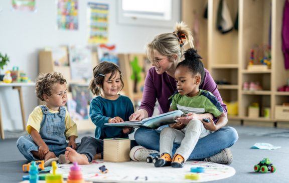 A group of children in a daycare or classroom setting engages with a teacher reading a book to them. The playroom background fosters an atmosphere of learning and playful interaction.