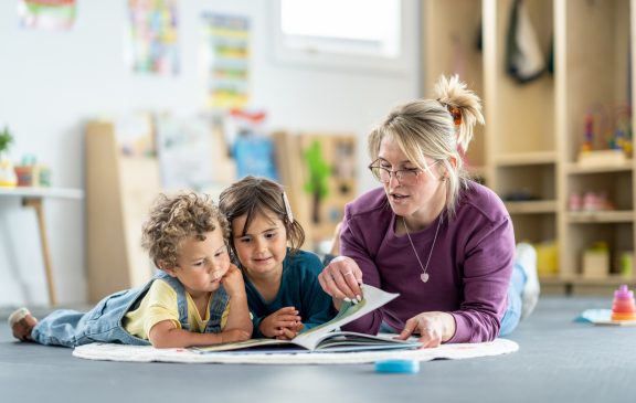 A teacher engaging with two children by reading together inside a well-lit playroom environment.