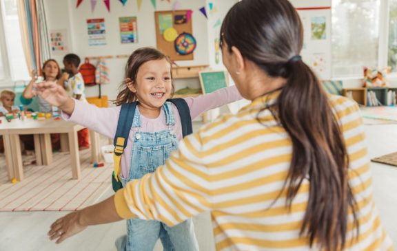 Preschool teacher in kindergarten classroom greeting new student, helping to easily assimilate with others.