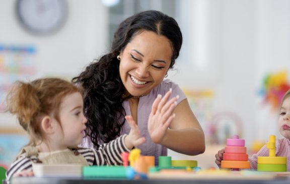 A smiling Black woman caregiver interacts with two young children at a sunny play table, guiding colorful stackable toys. Lively, supportive learning scene emphasizes care, teamwork, and early childhood education.