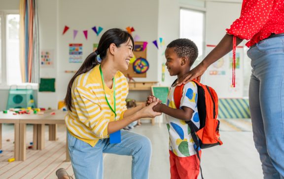 Preschool teacher in kindergarten classroom greeting new student, helping to easily assimilate with others.